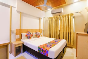 A hotel room with a bed, a nightstand, a desk, an air conditioning unit, and a curtain.