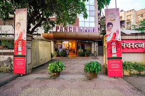 The entrance of a building with a sign reading "PANCHRATNA." There are two large red banners with text and images of a product. The entrance has a tiled pavement and potted plants.