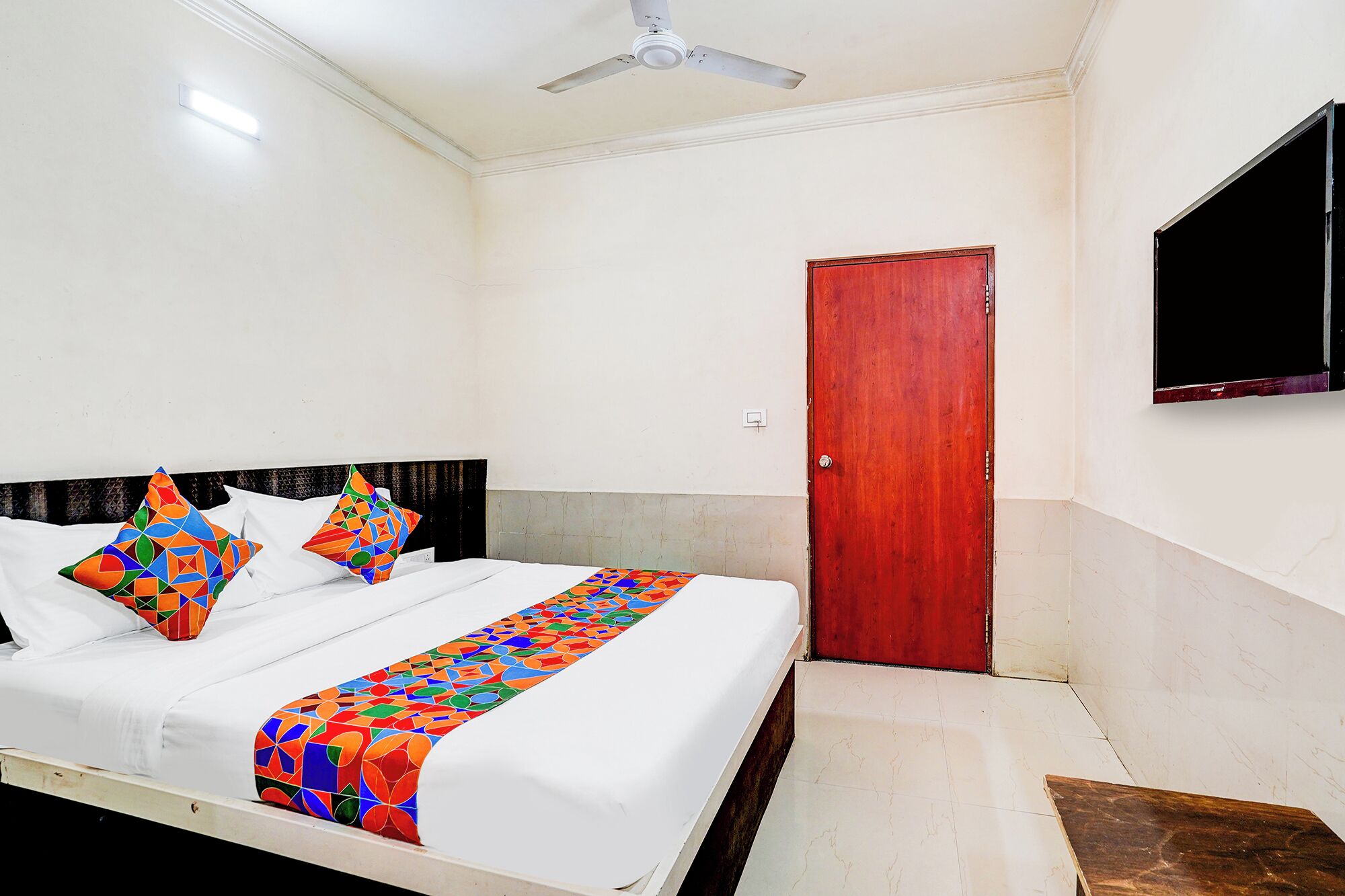 A hotel room with a bed, a ceiling fan, a wooden door, and a TV mounted on the wall.