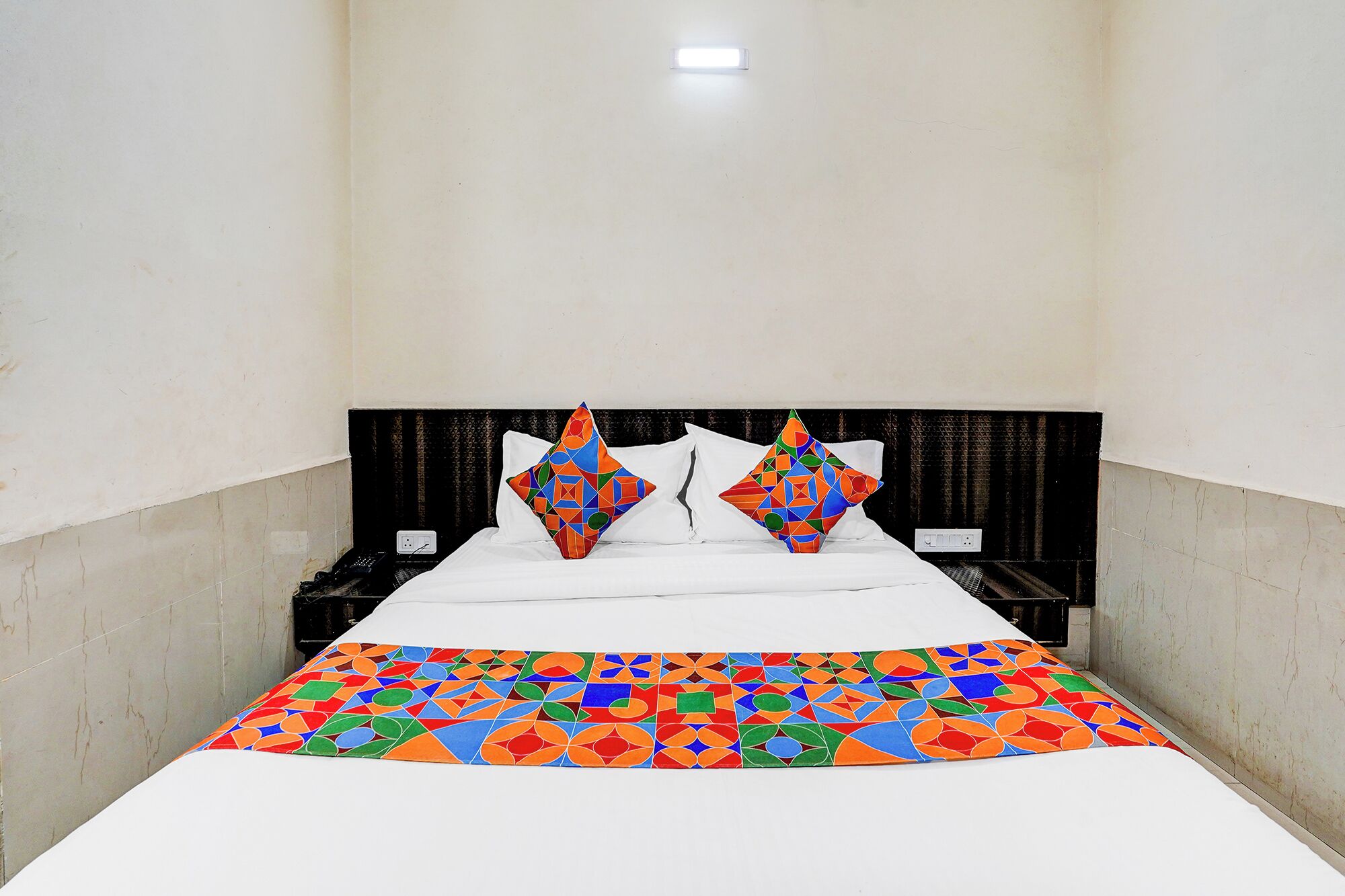 A single bed with a colorful geometric-patterned bedspread, two decorative pillows, a plain headboard, and a single bedside table with a phone.