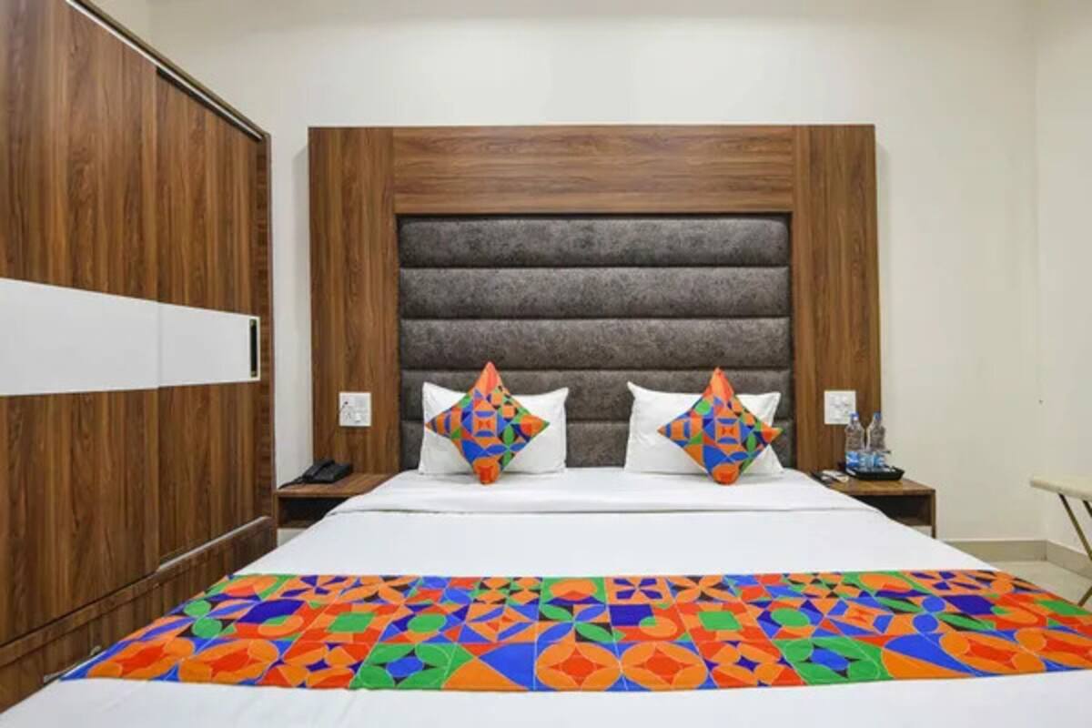 A hotel room with a wooden wardrobe, a bed with a colorful geometric-patterned bedspread, and a headboard with horizontal panels.