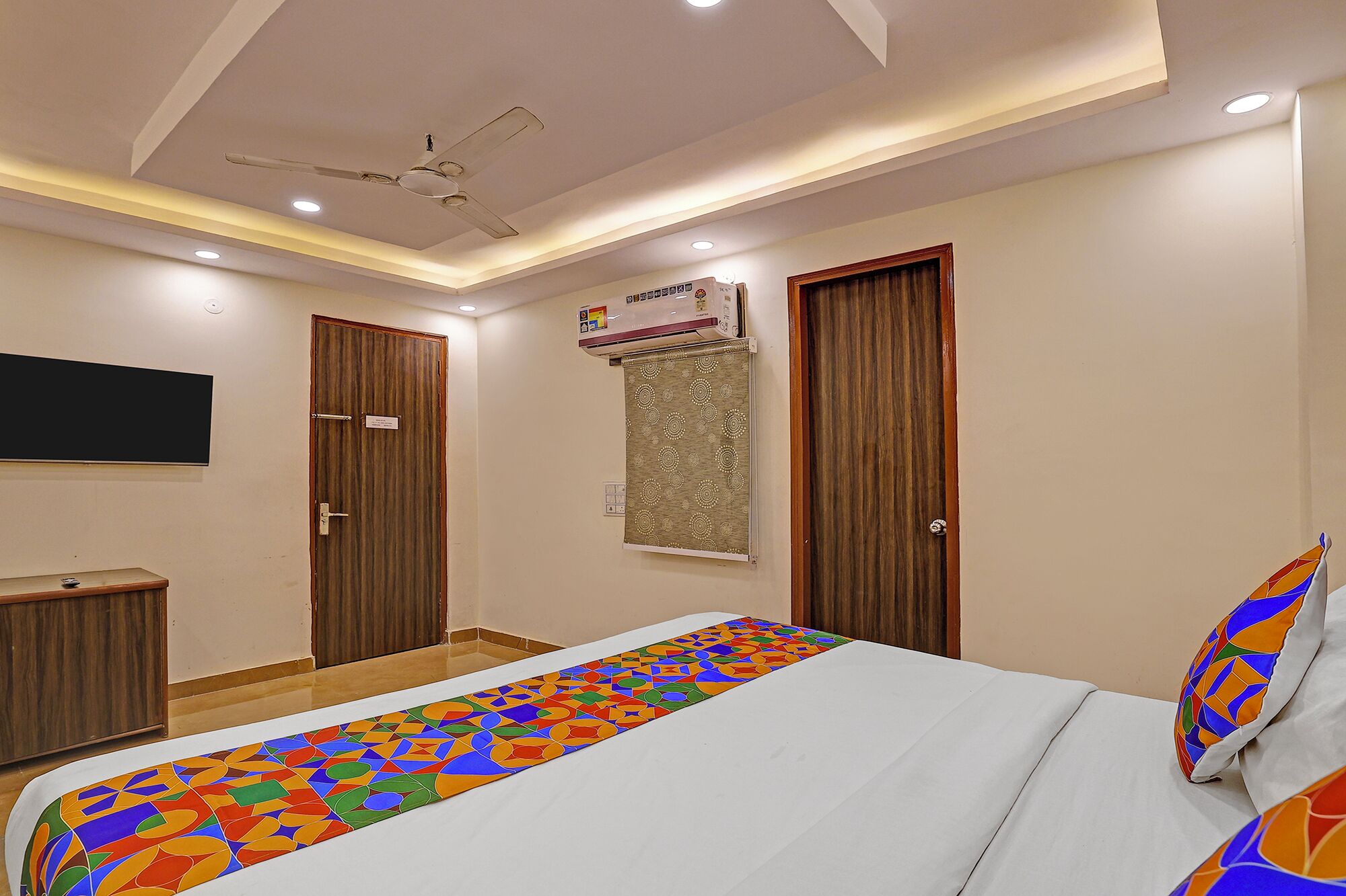 A hotel room with a bed, a ceiling fan, a mounted air conditioner, and a television on the wall.