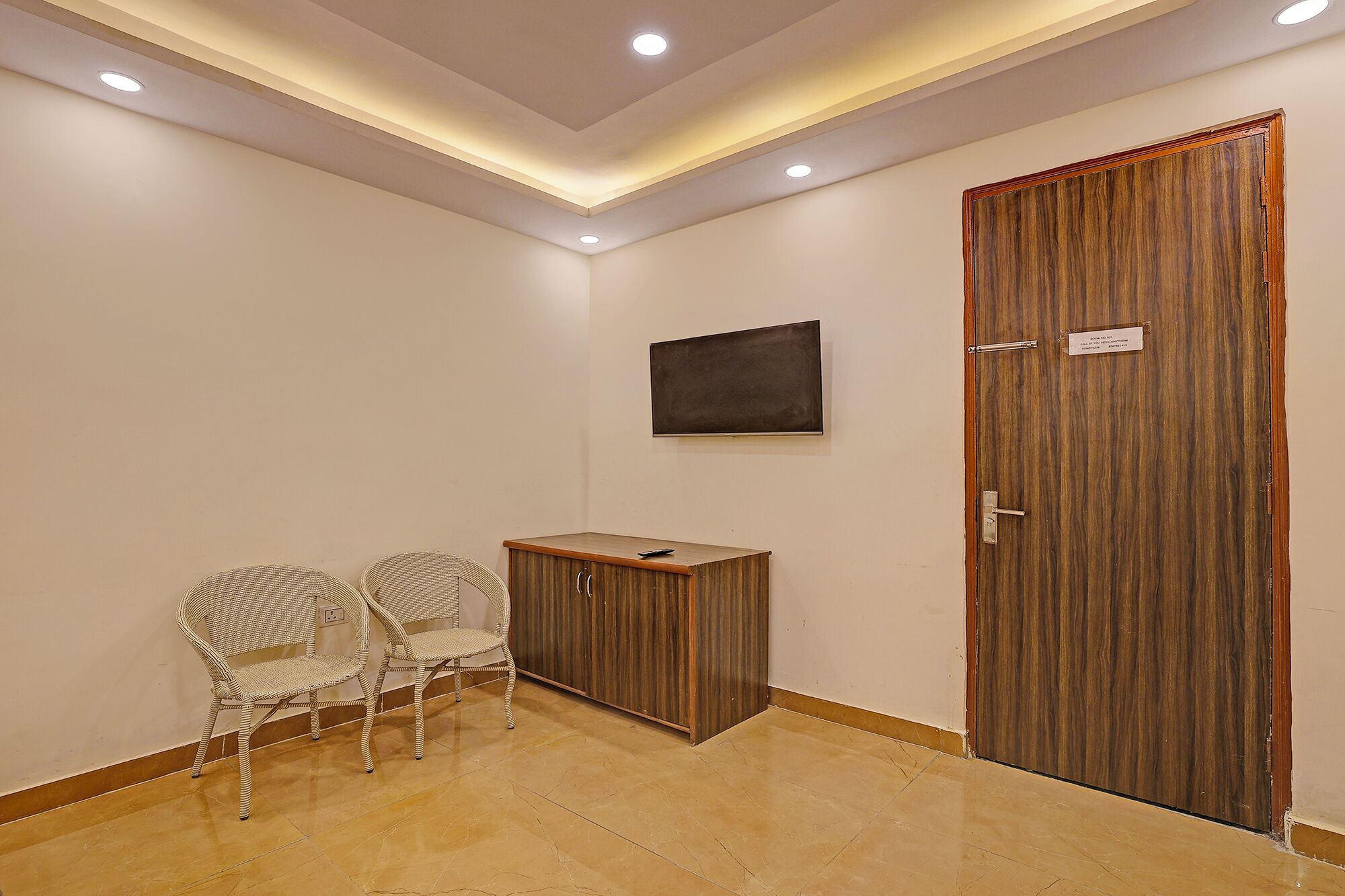 A room with a wooden door, a small cabinet, two wicker chairs, and a mounted flat-screen TV.
