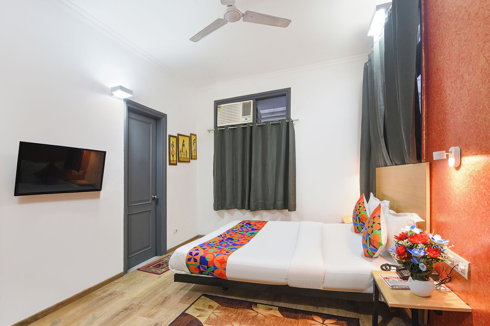 A hotel room with a bed, a ceiling fan, a TV mounted on the wall, and a window with curtains.