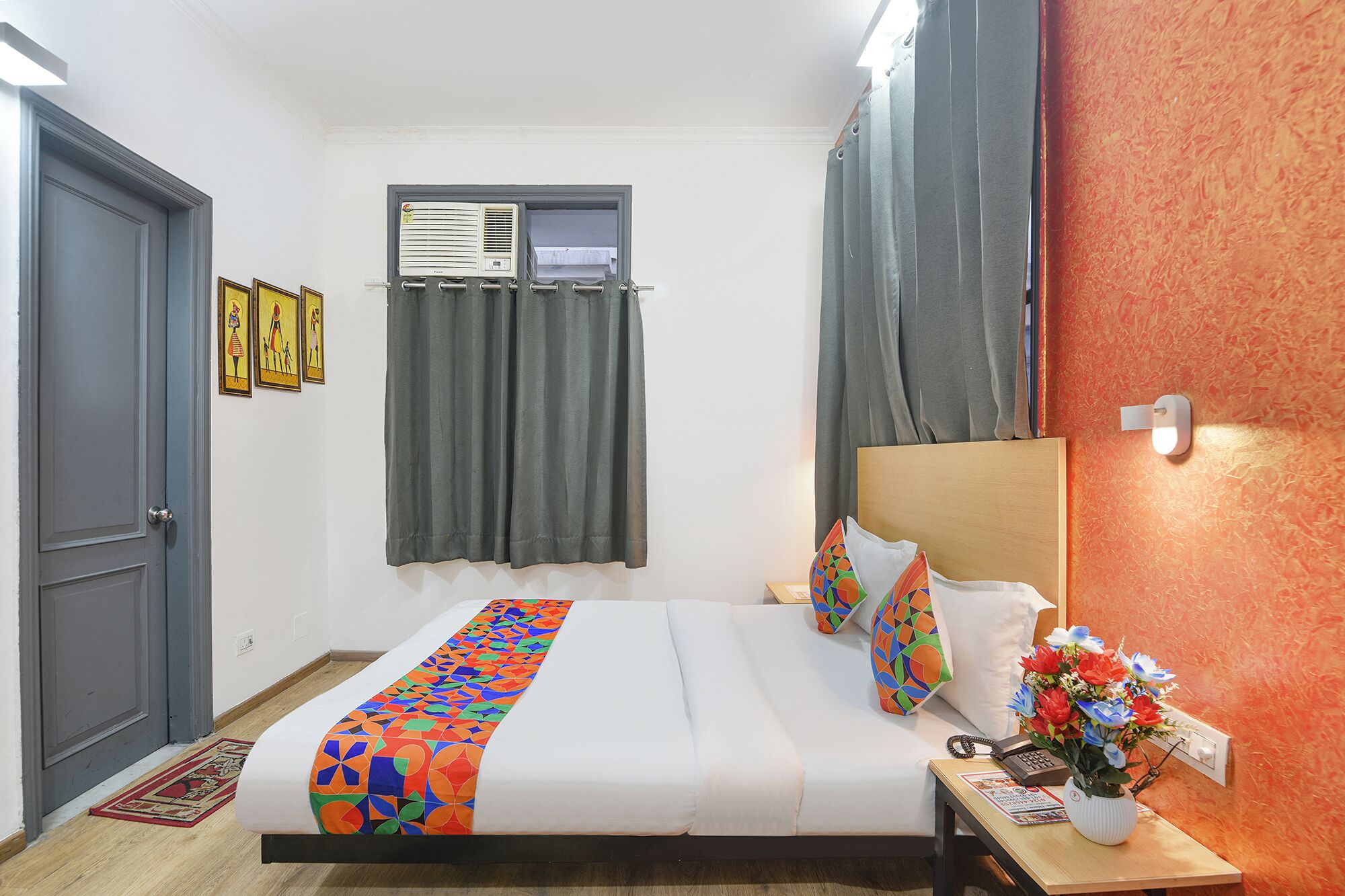 A hotel room with a bed, a bedside table with a vase of flowers, a wall-mounted air conditioner, and a window with curtains.