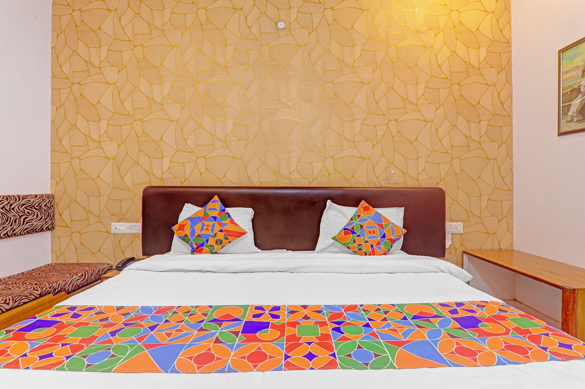 A hotel room with a patterned wall, a bed with a colorful bedspread, two pillows with geometric designs, and a nightstand with a lamp.