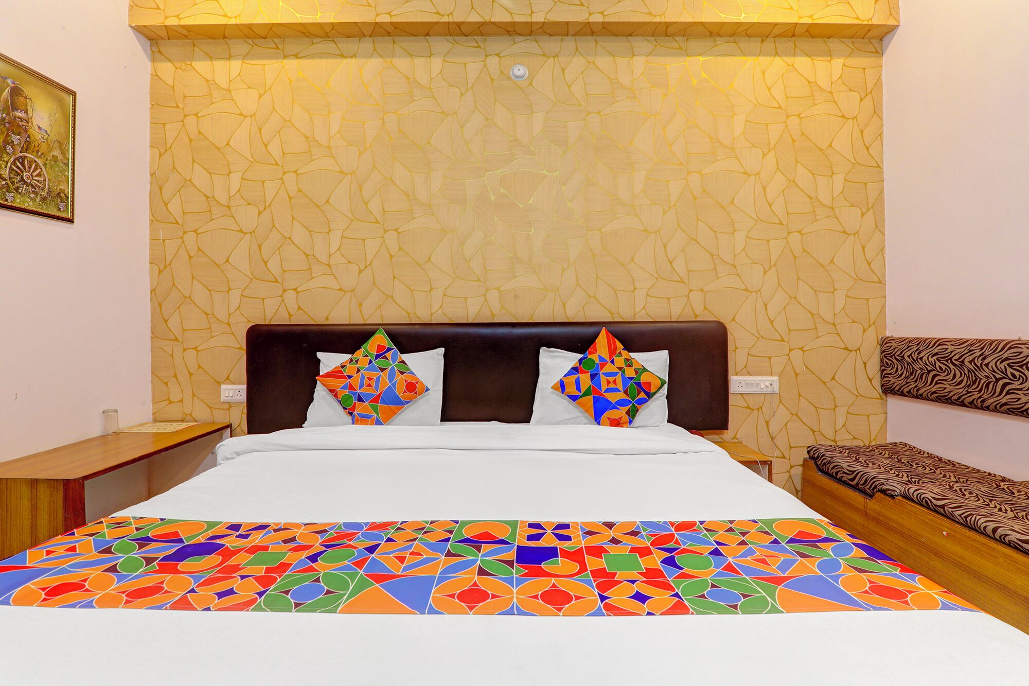 Une chambre d’hôtel avec un mur orné d’un motif, un lit recouvert d’une courtepointe géométrique aux couleurs vives, deux coussins décoratifs, une table de chevet et un banc en bois.