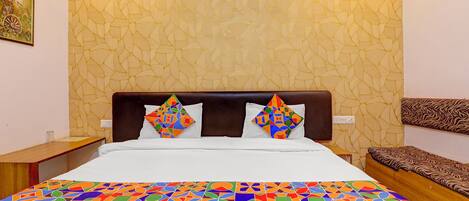 Une chambre d’hôtel avec un mur orné d’un motif, un lit recouvert d’une courtepointe géométrique aux couleurs vives, deux coussins décoratifs, une table de chevet et un banc en bois.