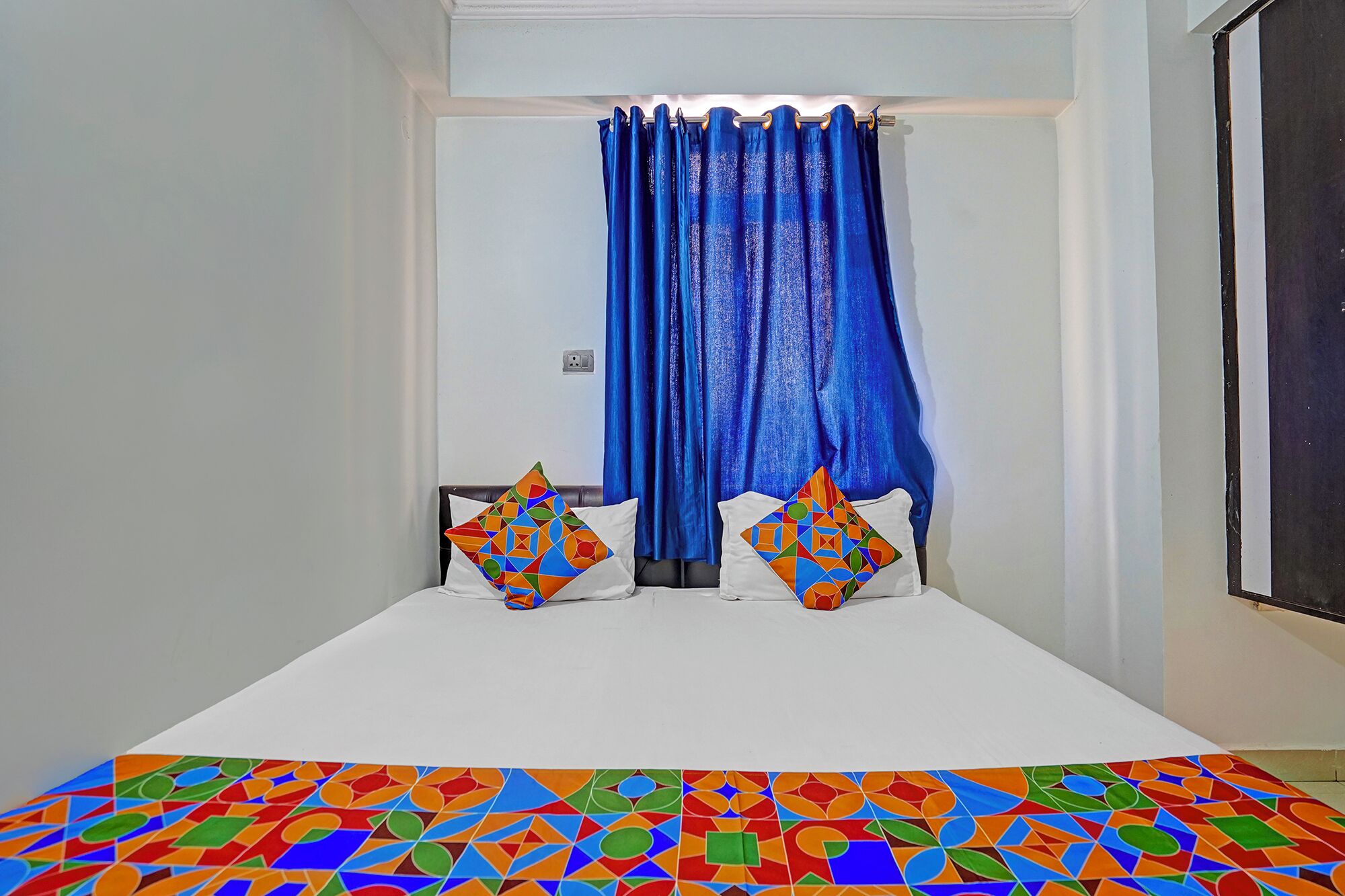 Quarto de hotel com uma cama branca, duas almofadas coloridas com padrão geométrico, uma cortina azul e piso estampado.