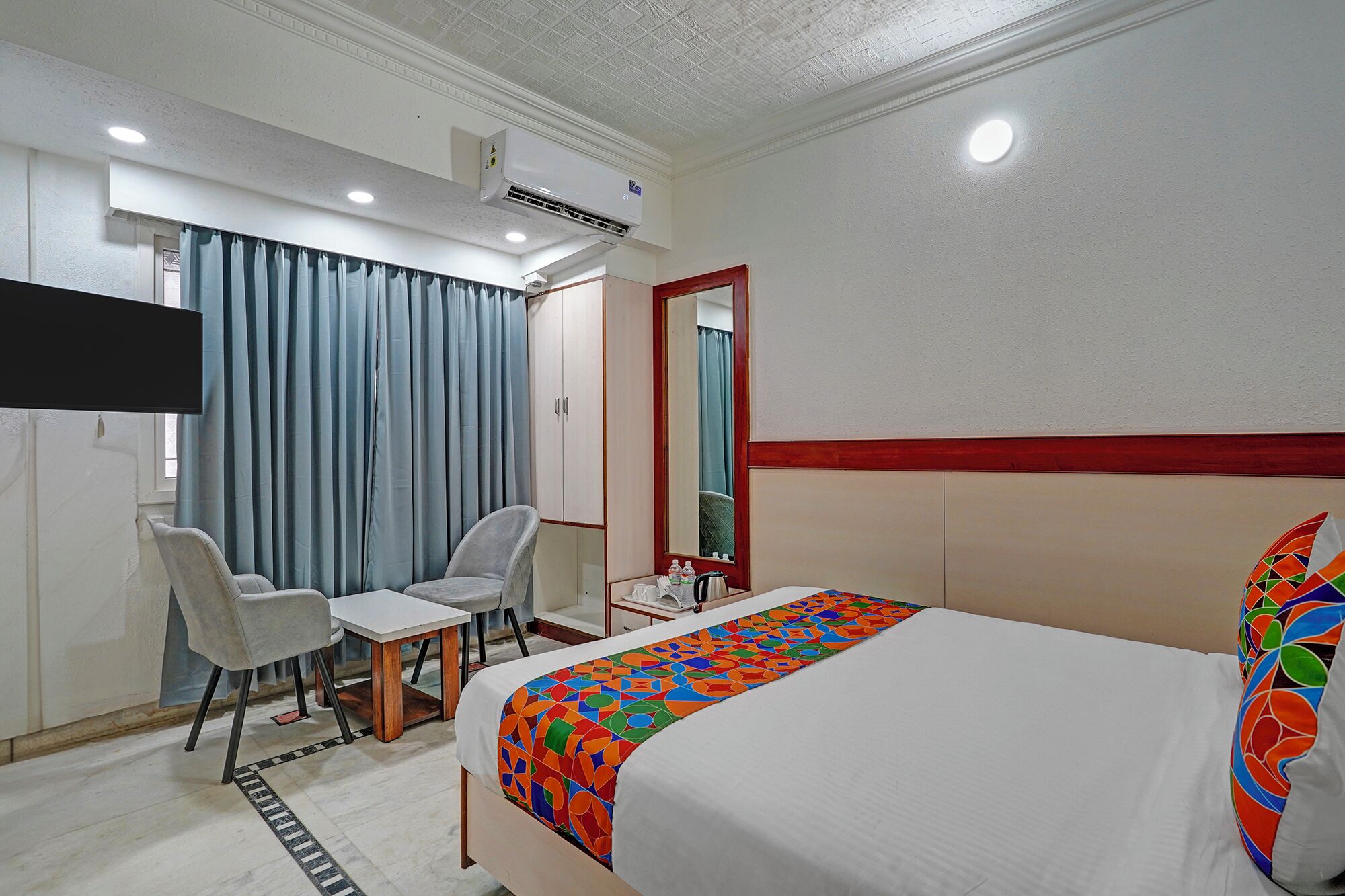 A hotel room with a bed, a desk, two chairs, a mirror, and an air conditioning unit.