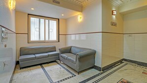 Ett hotellrum med en soffa, ett litet bord, ett fönster med persienner, ett kaklat golv med en dekorativ kant och en väggmonterad lampa.