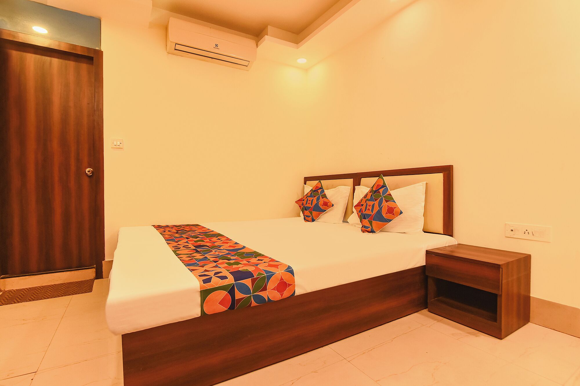 A bedroom with a wooden bed, a nightstand, a patterned bedspread, and an air conditioning unit on the wall.
