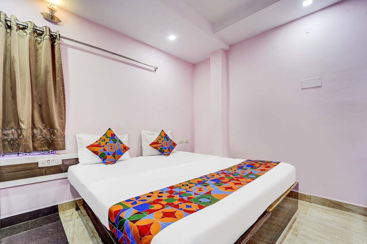 A hotel room with a bed, a nightstand, a lamp, a window with curtains, and a wall-mounted light fixture.