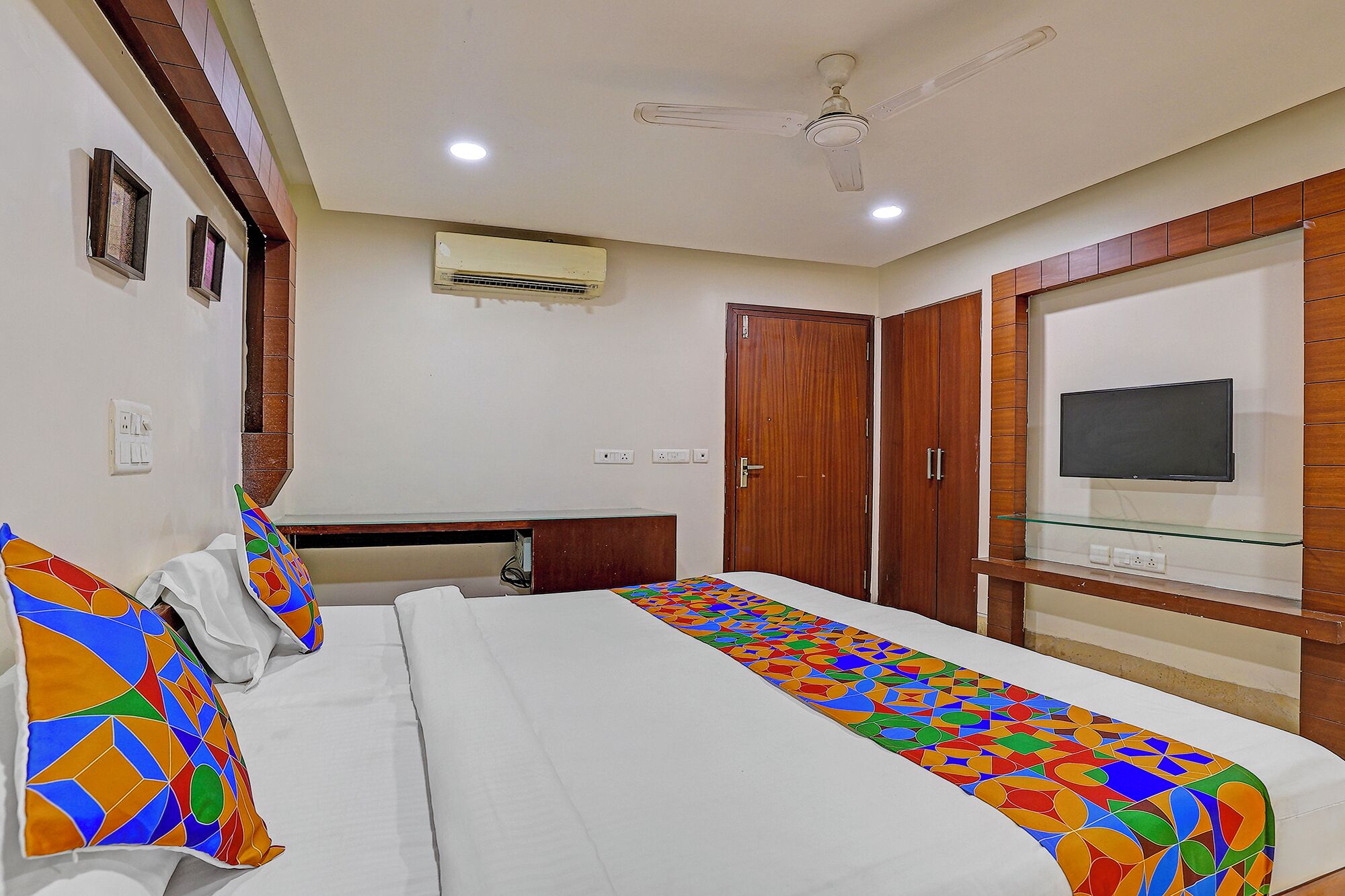 A hotel room with a bed, a TV mounted on the wall, a ceiling fan, and an air conditioning unit.