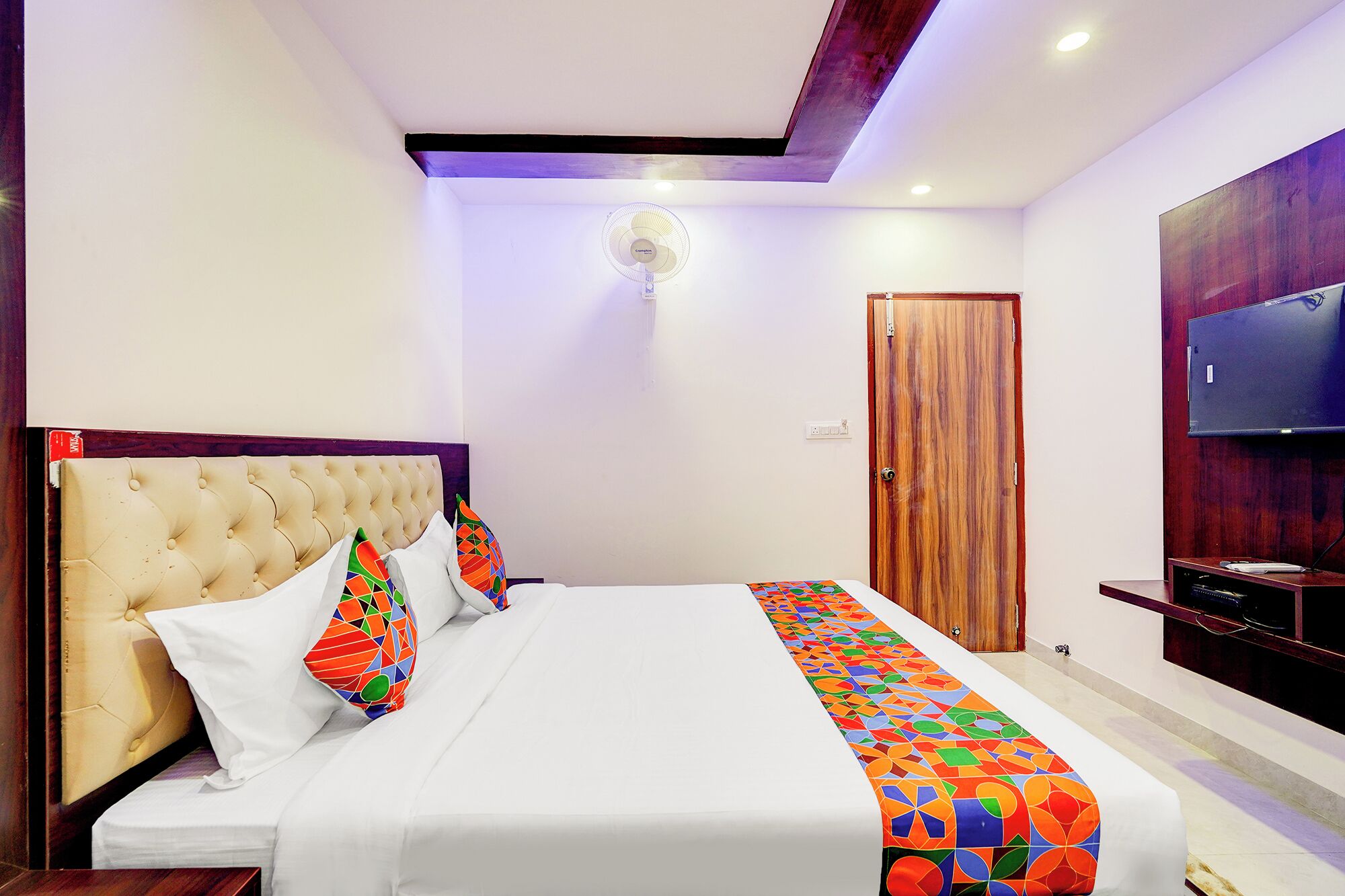 A modern hotel room with a tufted headboard, a large bed with colorful geometric-patterned pillows, a ceiling fan, a wooden door, and a flat-screen TV mounted on the wall.