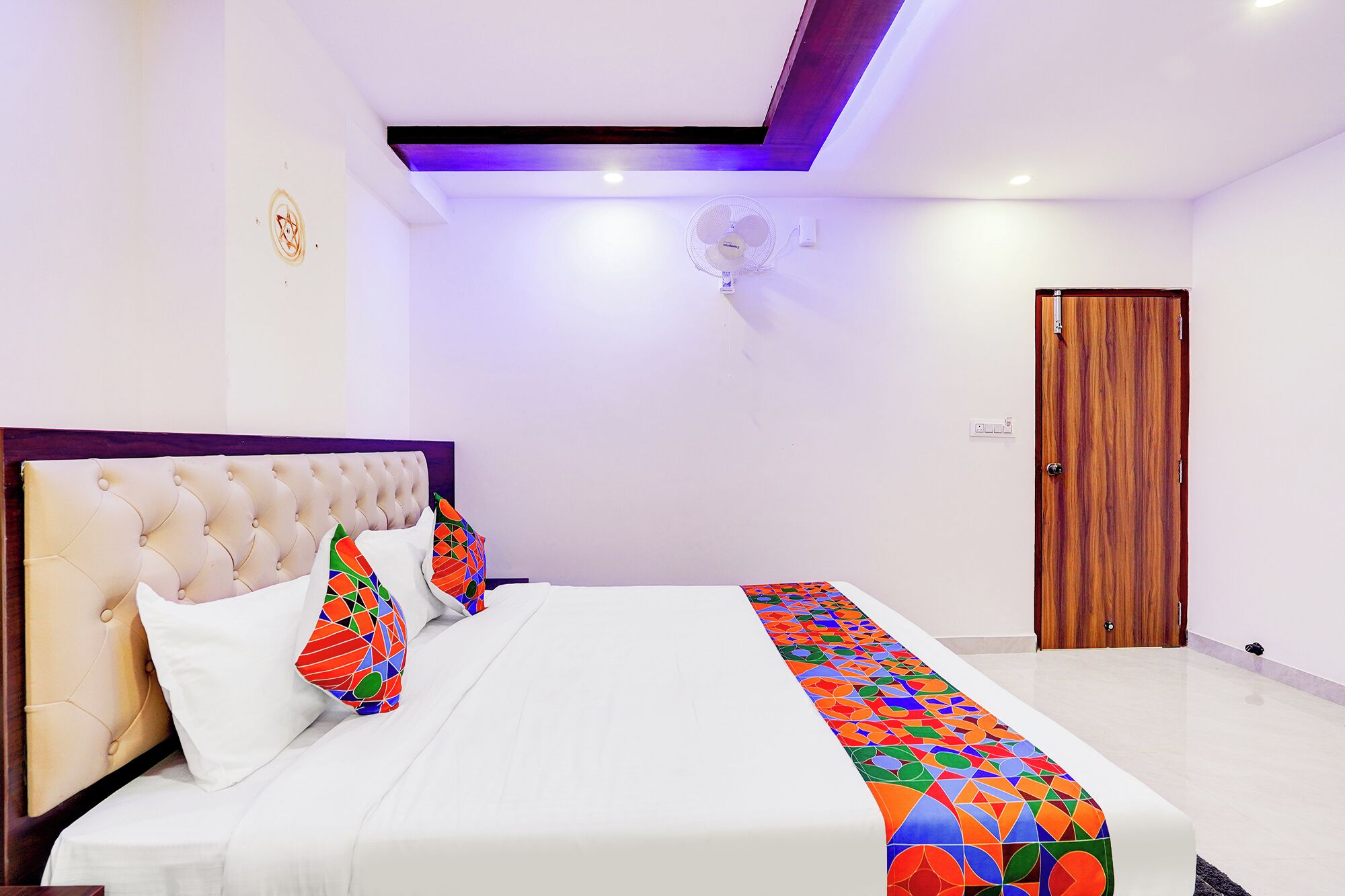Un dormitorio con cabecera capitonada, cama con colcha de diseño geométrico y colores vivos, un ventilador de pie y una puerta de madera.