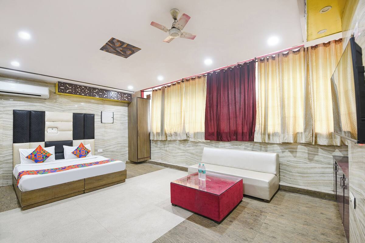 A hotel room with a bed, a sofa, a glass coffee table, curtains, a ceiling fan, and an air conditioning unit.