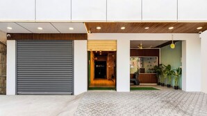 Una entrada de edificio moderna con una puerta persiana cerrada, una zona de recepción con mostrador y plantas, y un espacio interior visible con techo de madera e iluminación empotrada.