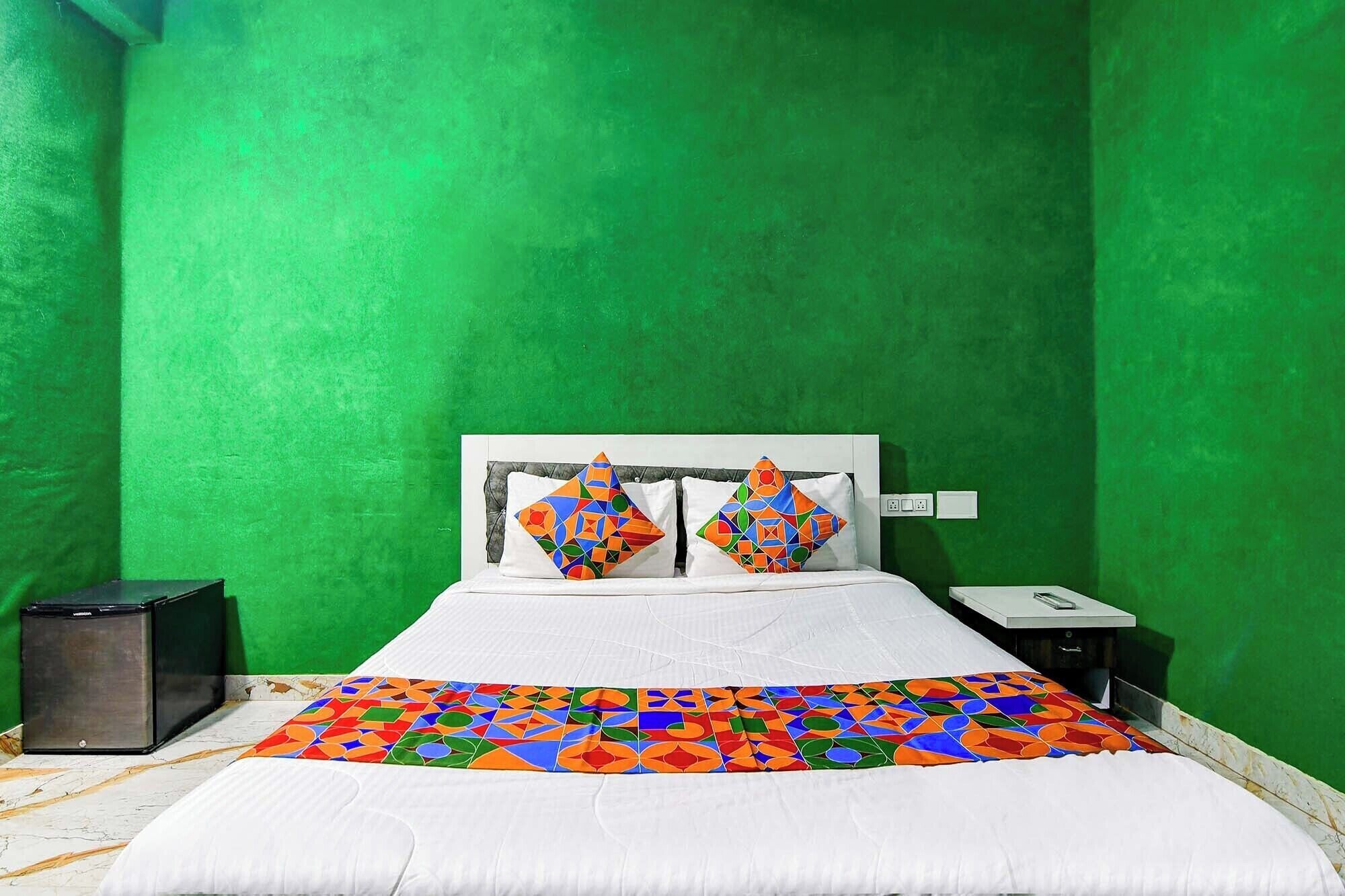 Quarto com parede verde, cama com colcha de padrão geométrico colorido, duas almofadas com padrão semelhante, cabeceira branca e mesa de cabeceira com um telefone e um pequeno alto-falante preto.