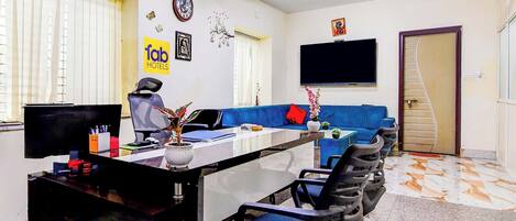 A hotel room with a desk, chairs, a sofa, a flat-screen TV, a clock, and a ceiling fan.
