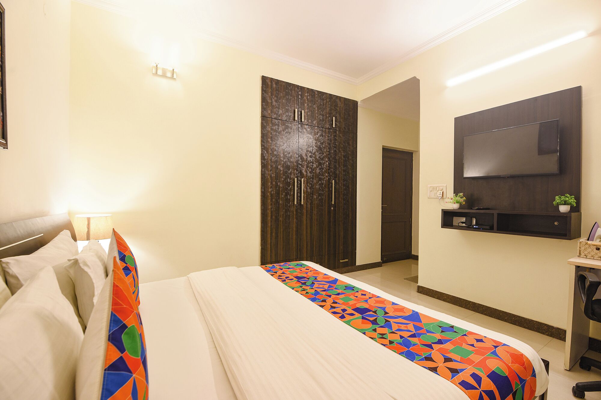 A hotel room with a bed, a wardrobe, a TV mounted on the wall, and a small shelf with a plant.