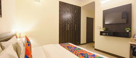 A hotel room with a bed, a wardrobe, a TV mounted on the wall, and a small shelf with a plant.