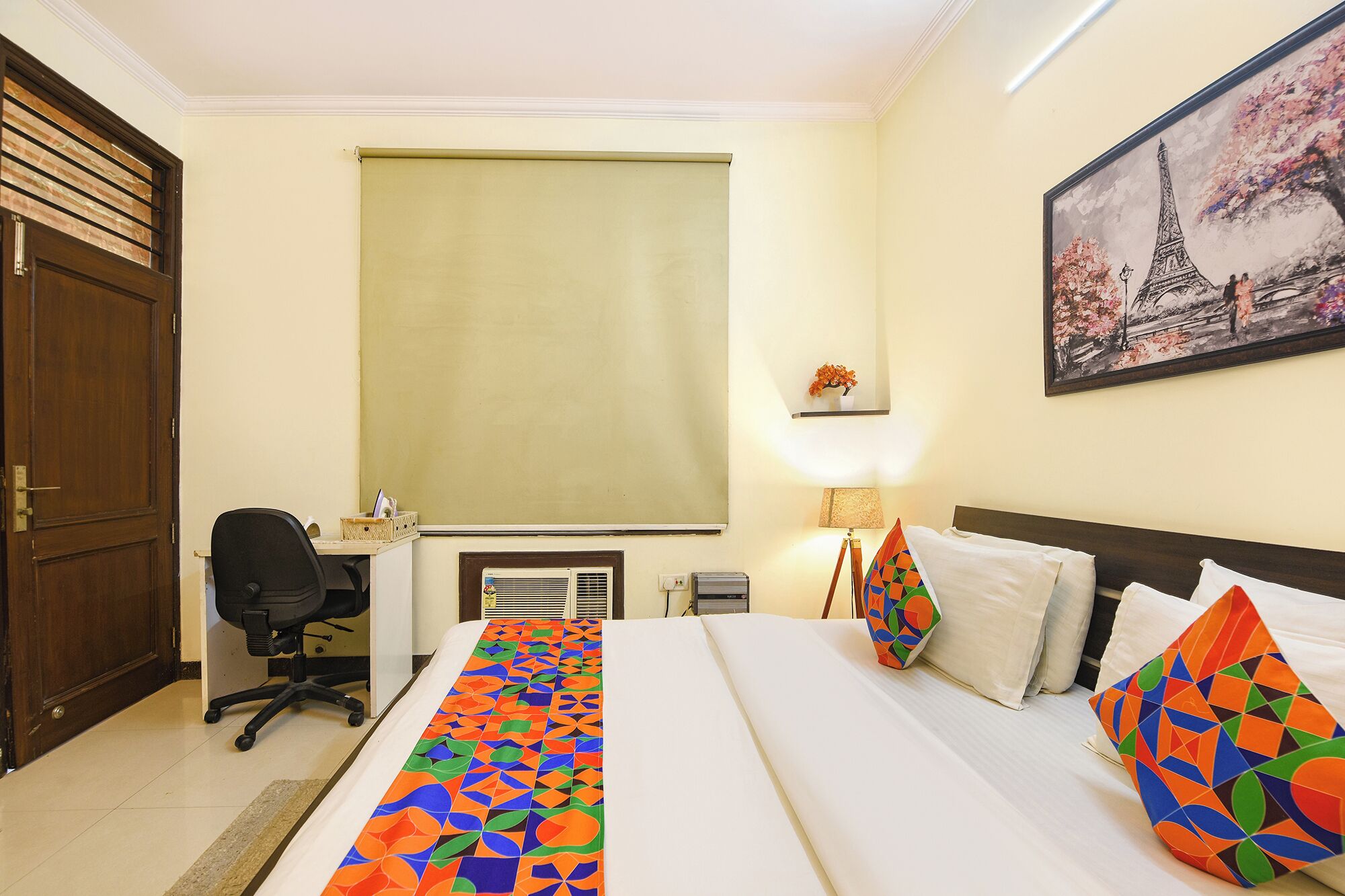 A hotel room with a bed, a desk, a chair, a window with a blind, a lamp, and a painting of the Eiffel Tower.