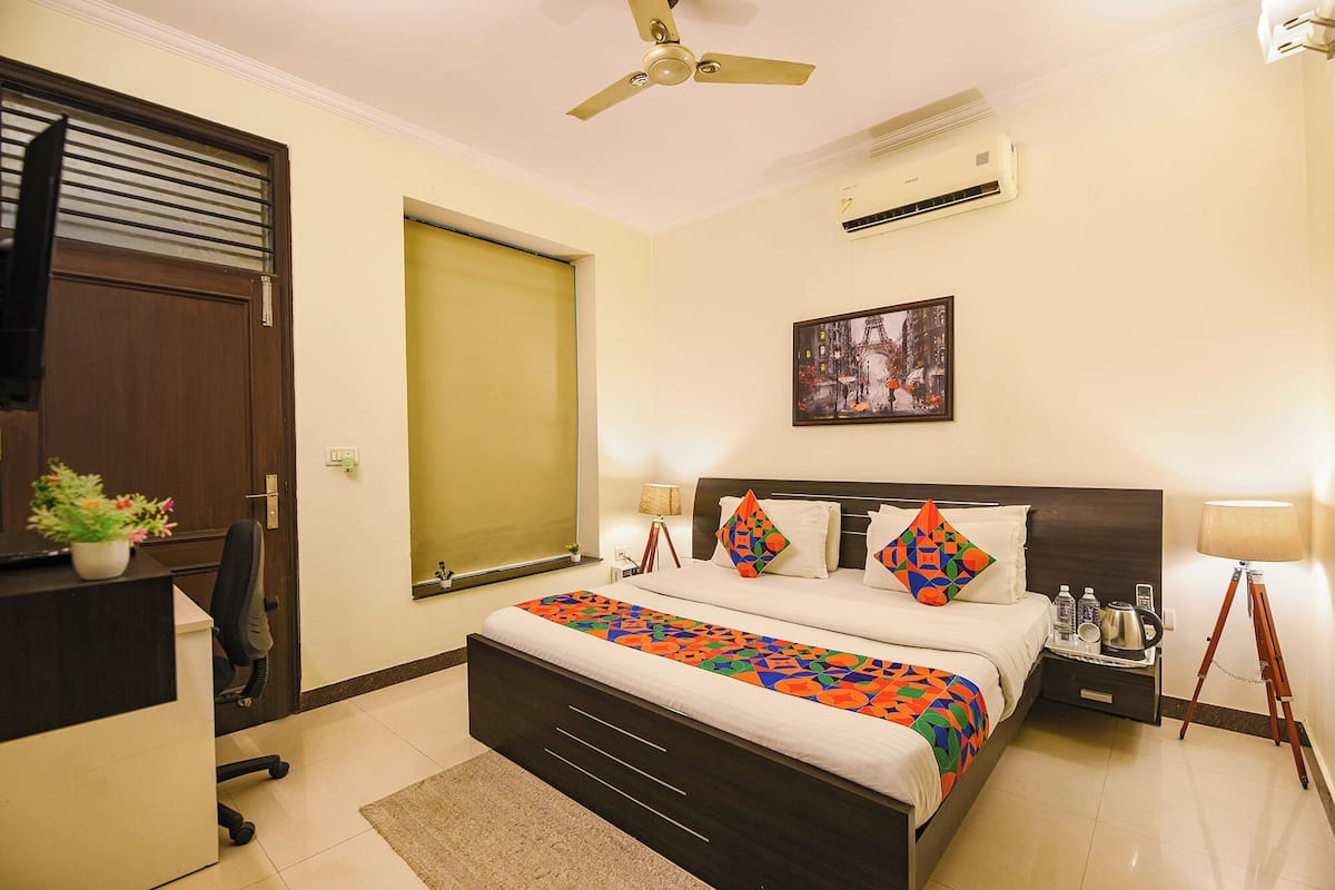 A hotel room with a bed, bedside tables, a desk, a chair, a ceiling fan, an air conditioning unit, a window with blinds, a framed picture on the wall, and a small potted plant.