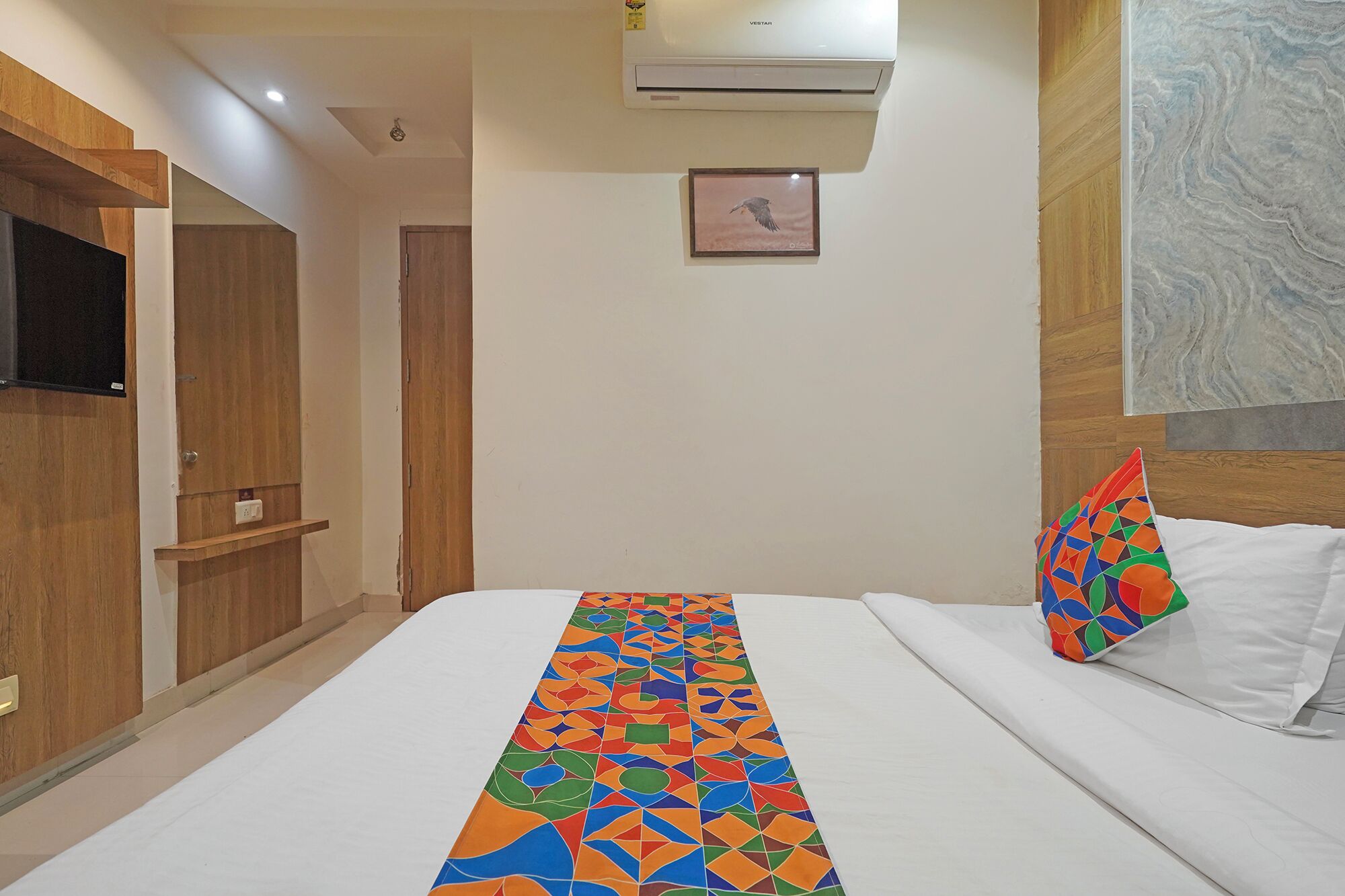 Habitación de hotel con un corredor largo y de estampado geométrico en colores vivos, cama con sábanas blancas y almohada colorida, televisor montado y un cuadro abstracto grande en la pared.