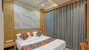 Une chambre d’hôtel avec un grand lit, une tête de lit en bois, un ventilateur de plafond et une vue sur l’extérieur à travers des rideaux gris.