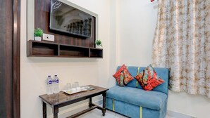 Ein Wohnzimmer mit einem blauen Sofa, zwei farbenfrohen Kissen, einem hölzernen Couchtisch, einem an einer Holzablage montierten Fernseher und einem gemusterten Vorhang.