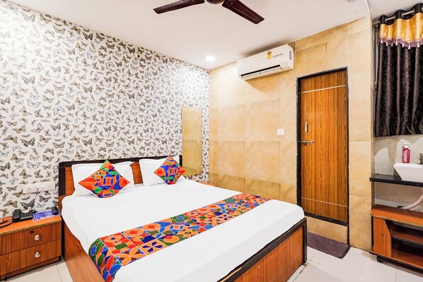 A bedroom with a bed, a nightstand, an air conditioning unit, a wooden door, and a bathroom with a sink and mirror. - Fabhotel Krishna Inn (Surat)