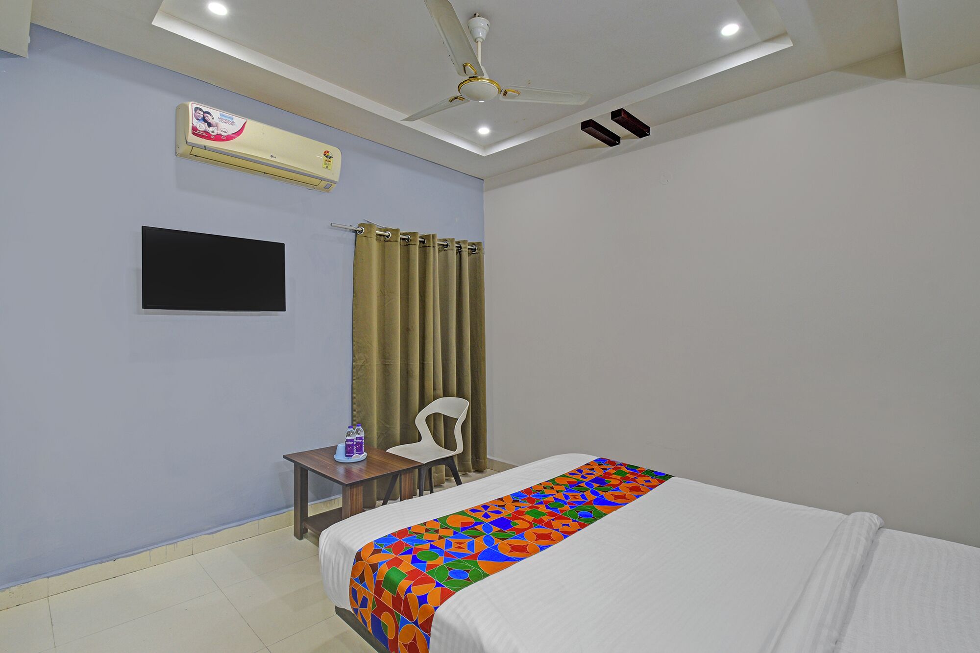 A hotel room with a bed, a desk, a chair, a ceiling fan, an air conditioning unit, and a flat-screen TV.