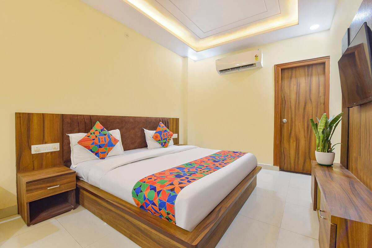 Una habitación de hotel moderna con un cabecero de madera, una cama con ropa de cama geométrica y colorida, una mesita de noche, una cómoda de madera, una planta en maceta y un armario de madera.
