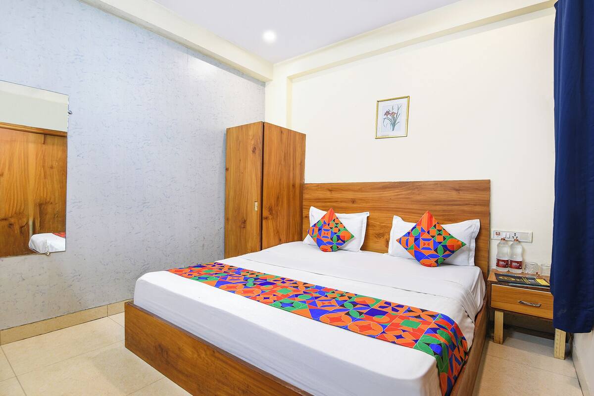A hotel room with a wooden bed, white bedding, colorful geometric pillows, a wooden wardrobe, a small bedside table with a lamp, and a framed picture on the wall.