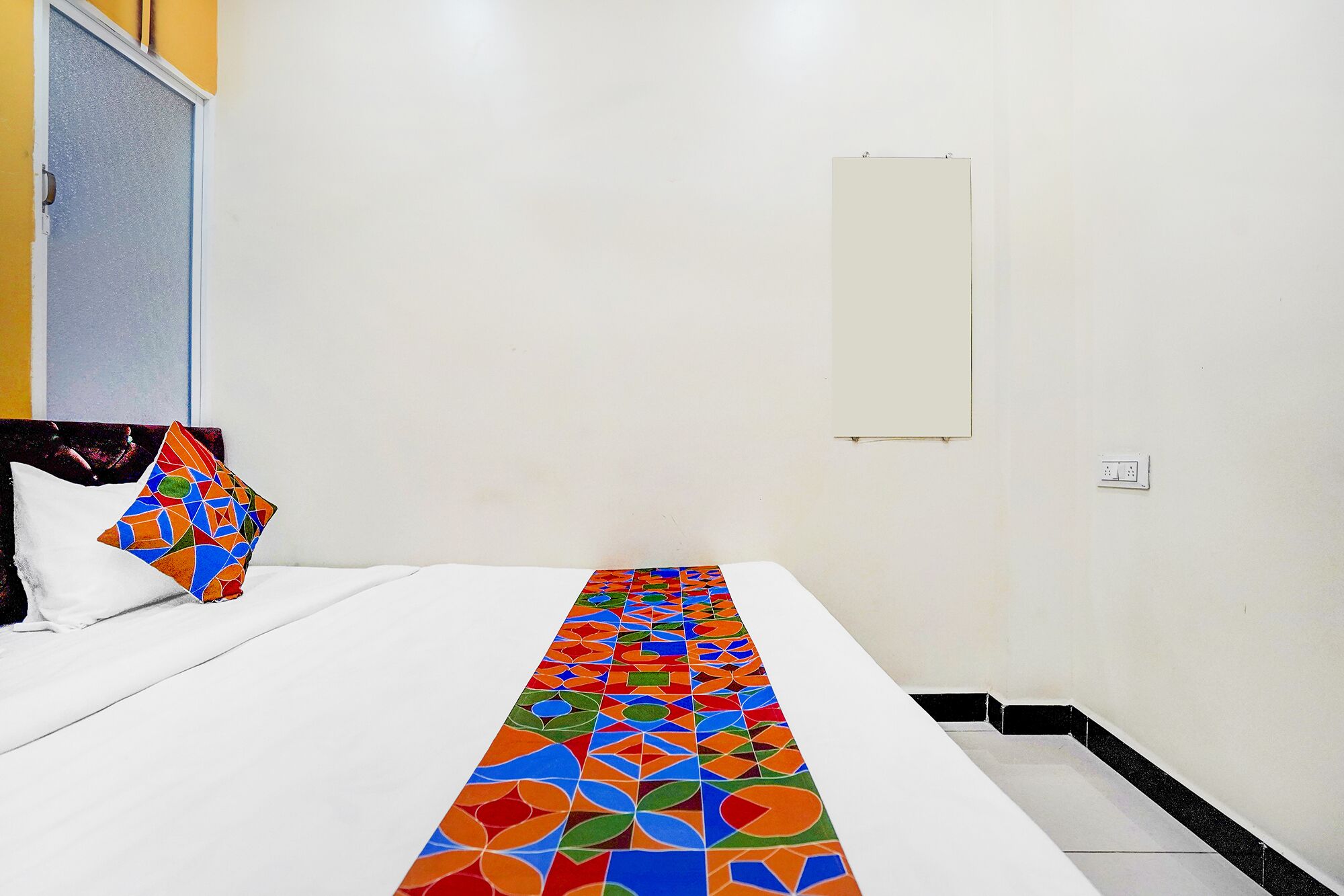 A hotel room with a bed, a colorful geometric-patterned runner, a plain wall, and a small whiteboard.