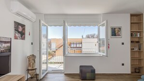 A modern living room with a large window, wooden flooring, a television, and a bookshelf. - Bells & Whistles, Comfortable Condo (Split)