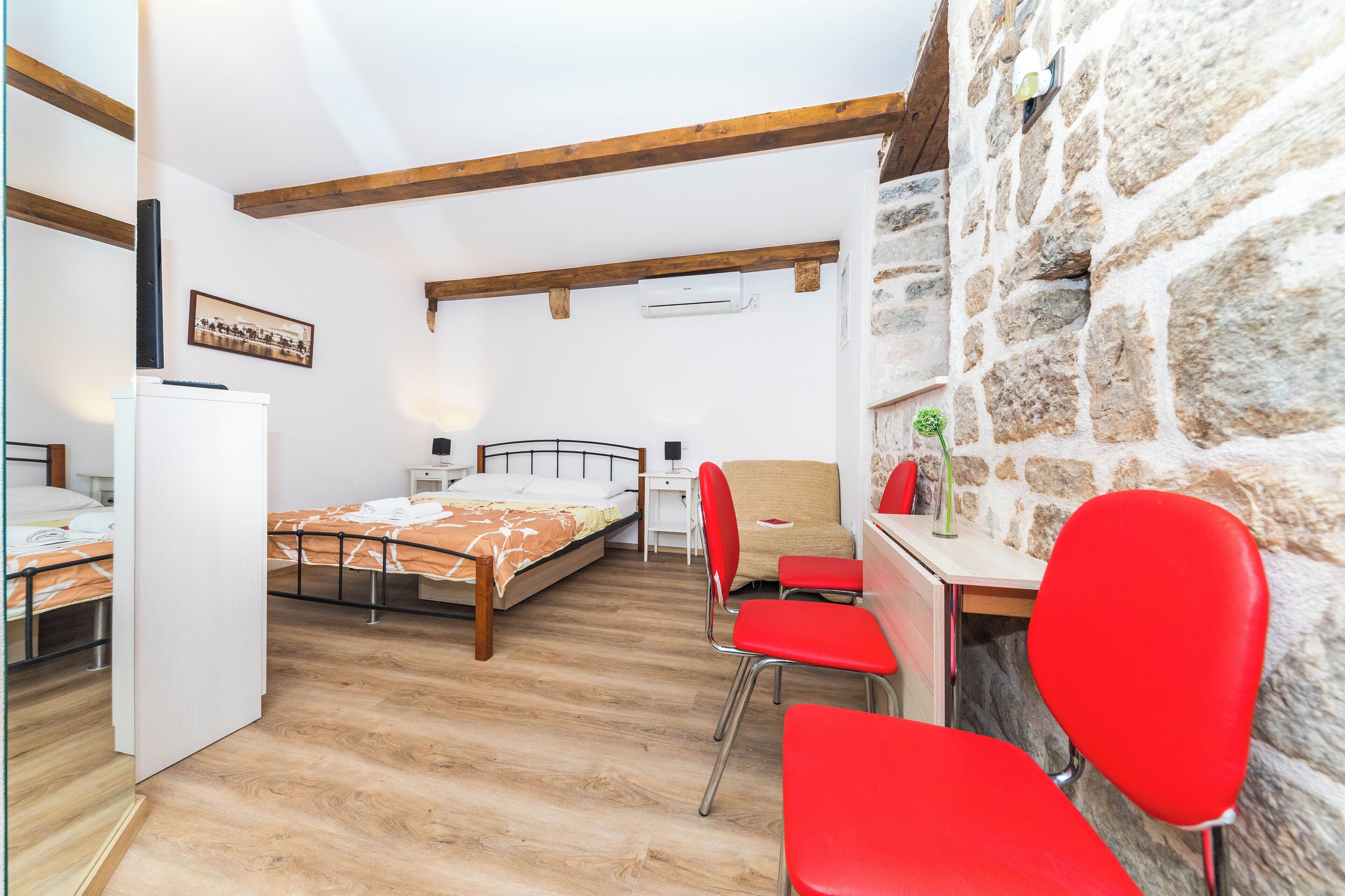A room with a bed, a desk, and a chair. There is a stone wall and wooden beams on the ceiling.