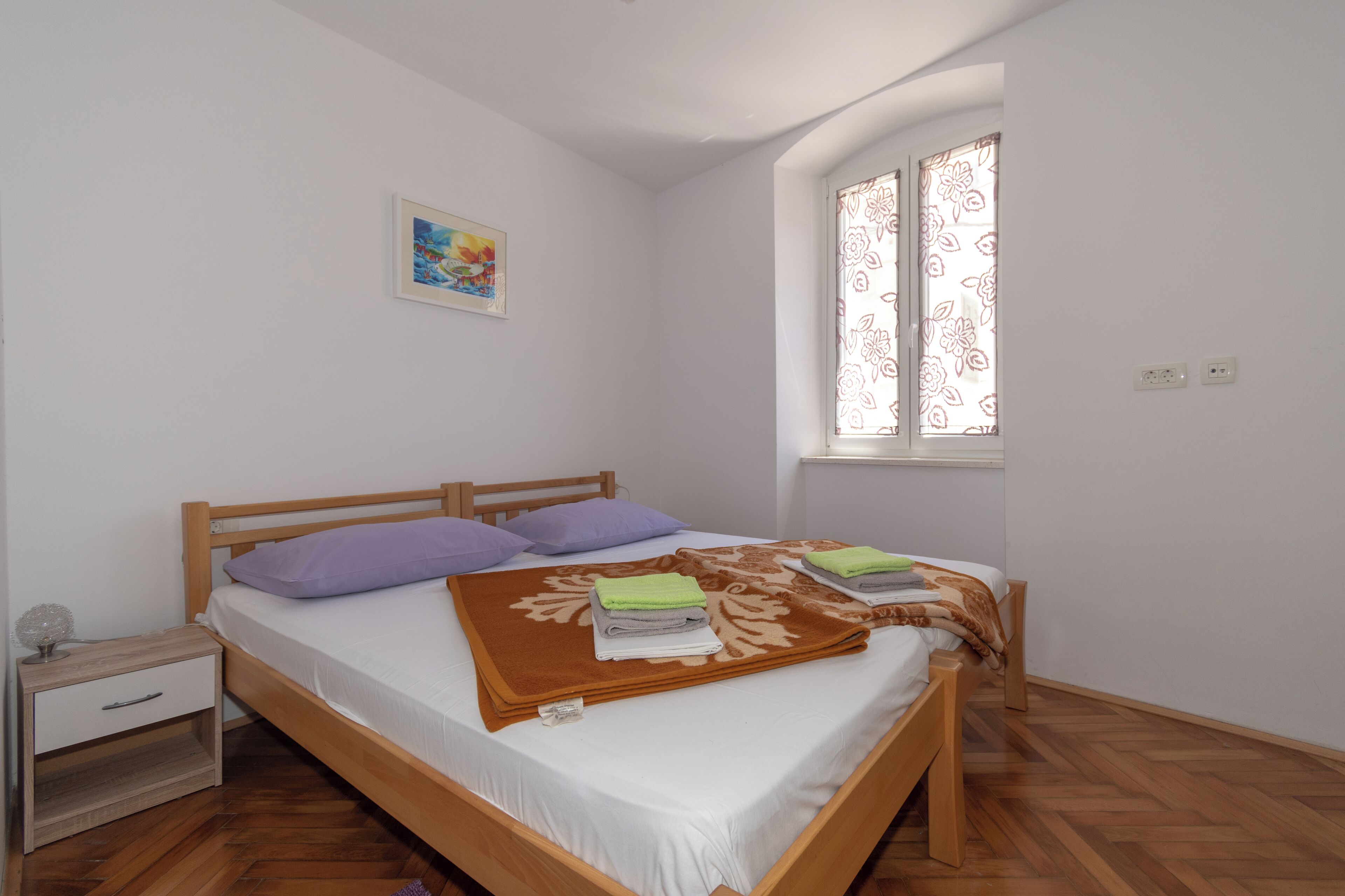 Un lit bien fait, recouvert d’une couverture marron, avec une tête de lit en bois et deux oreillers. On aperçoit une petite table de chevet avec une lampe et un tiroir. Une fenêtre ornée de rideaux à motifs floraux est visible, et un tableau est accroché au mur.