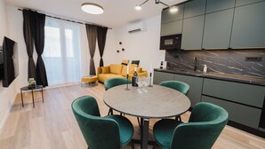 Egy modern nappali, melyben van egy sárga kanapé, egy kerek étkezőasztal zöld székekkel, valamint egy konyhasarok sötét színű szekrényekkel és márvány munkalappal.