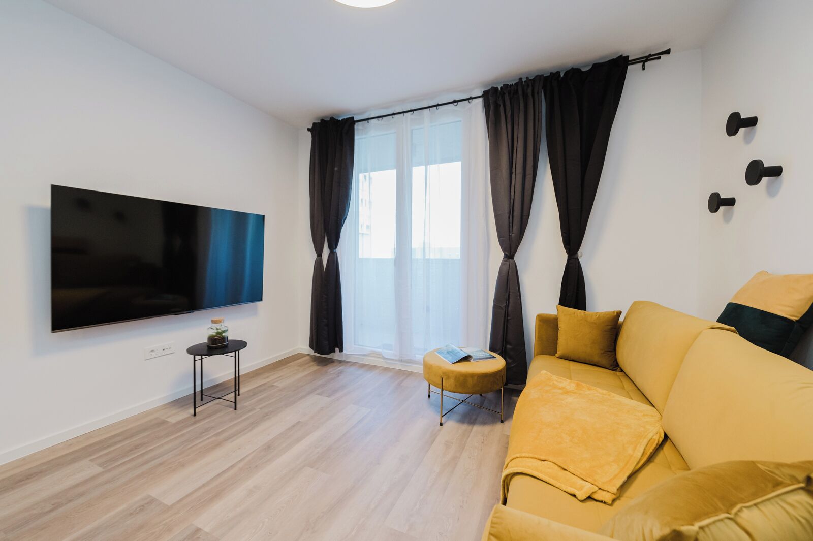 Un salon moderne avec un grand téléviseur à écran plat, un canapé jaune, un petit pouf rond et une table d’appoint.