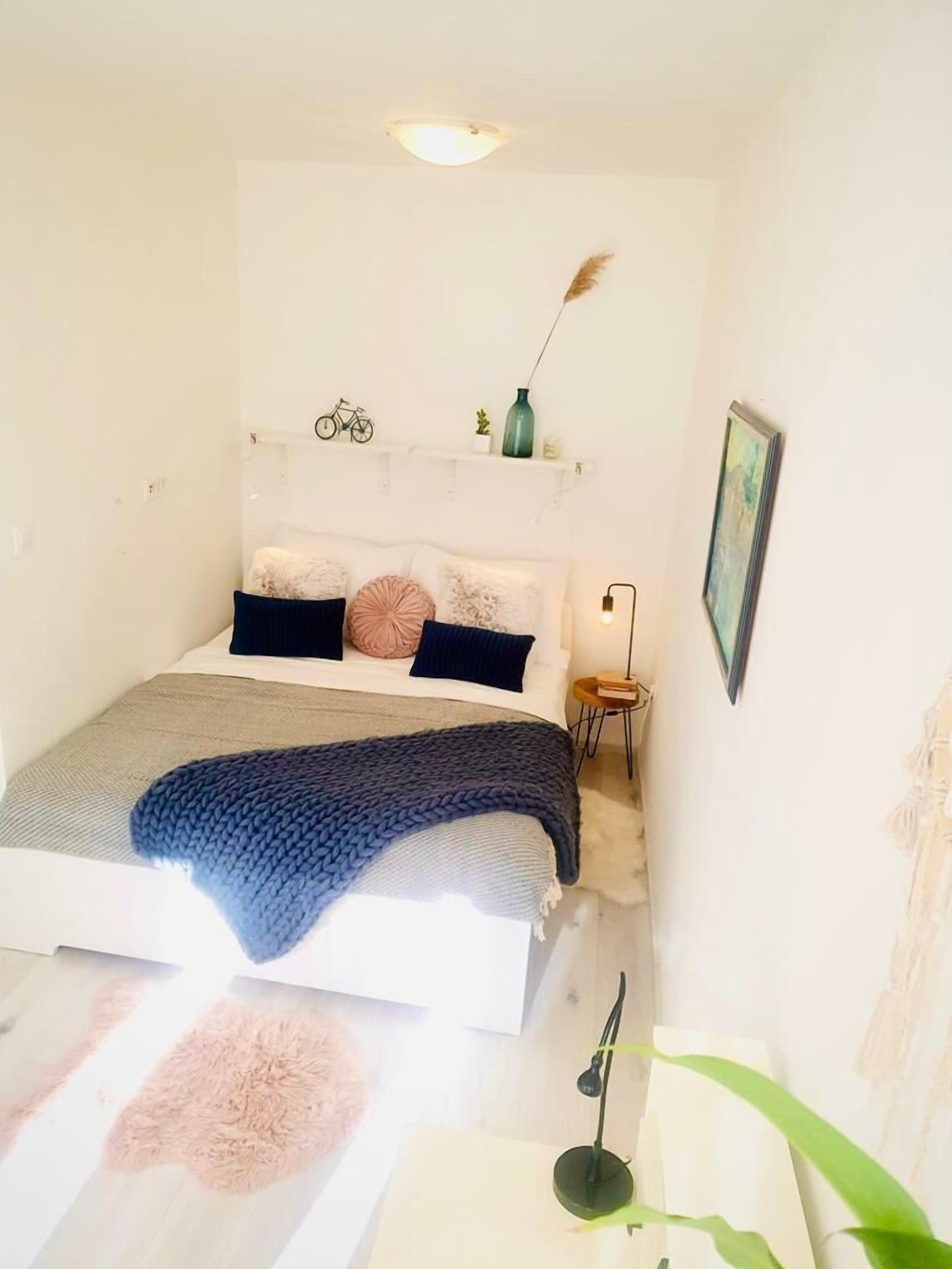 A bedroom with a bed, a bedside table, a lamp, a wall-mounted shelf with a bicycle, a plant, and a painting.