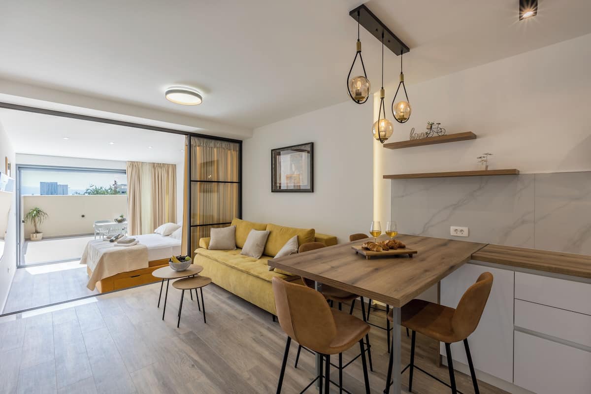 A modern hotel room with a wooden dining table, a sofa, and a balcony with a city view.