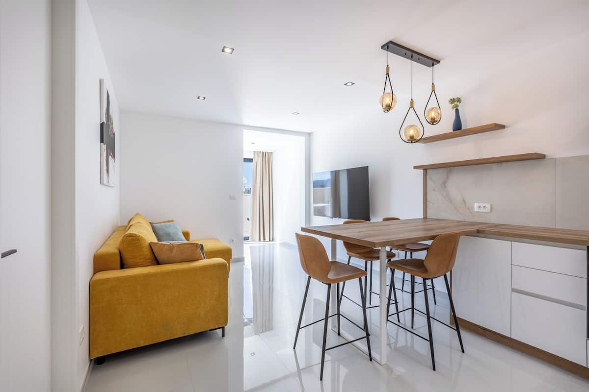 A modern kitchen with a wooden table and bar stools, a yellow sofa, and pendant lighting.