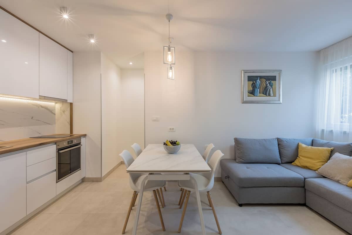A modern kitchen with white cabinets and a built-in oven, a dining table with chairs, and a gray sofa with yellow pillows.