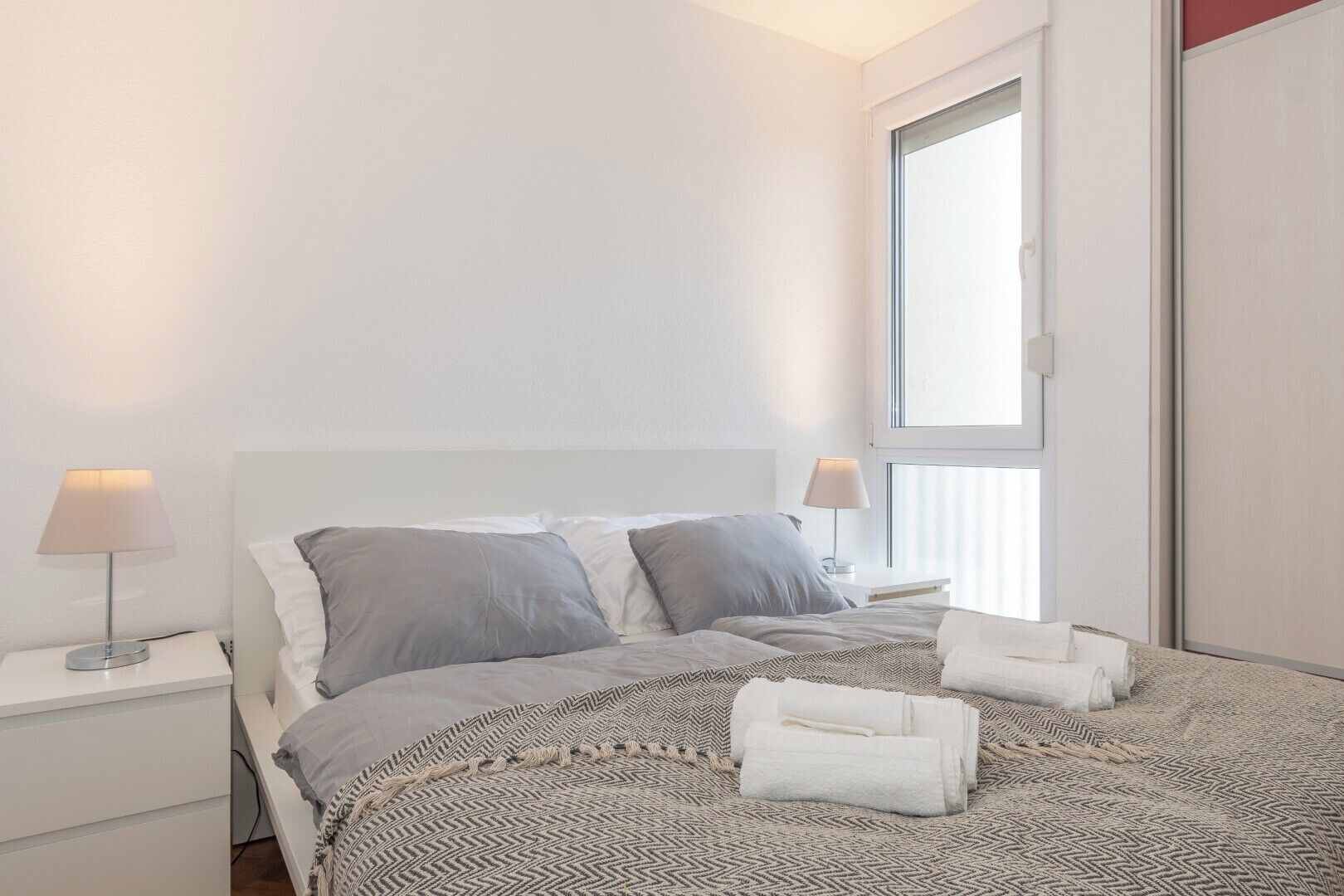 A neatly made bed with grey and white bedding, two bedside lamps, and a window with blinds.