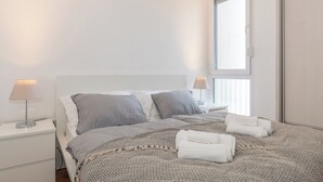 A neatly made bed with grey and white bedding, two bedside lamps, and a window with blinds. - Zozo, Modern Two-Bedroom Suite (Split)