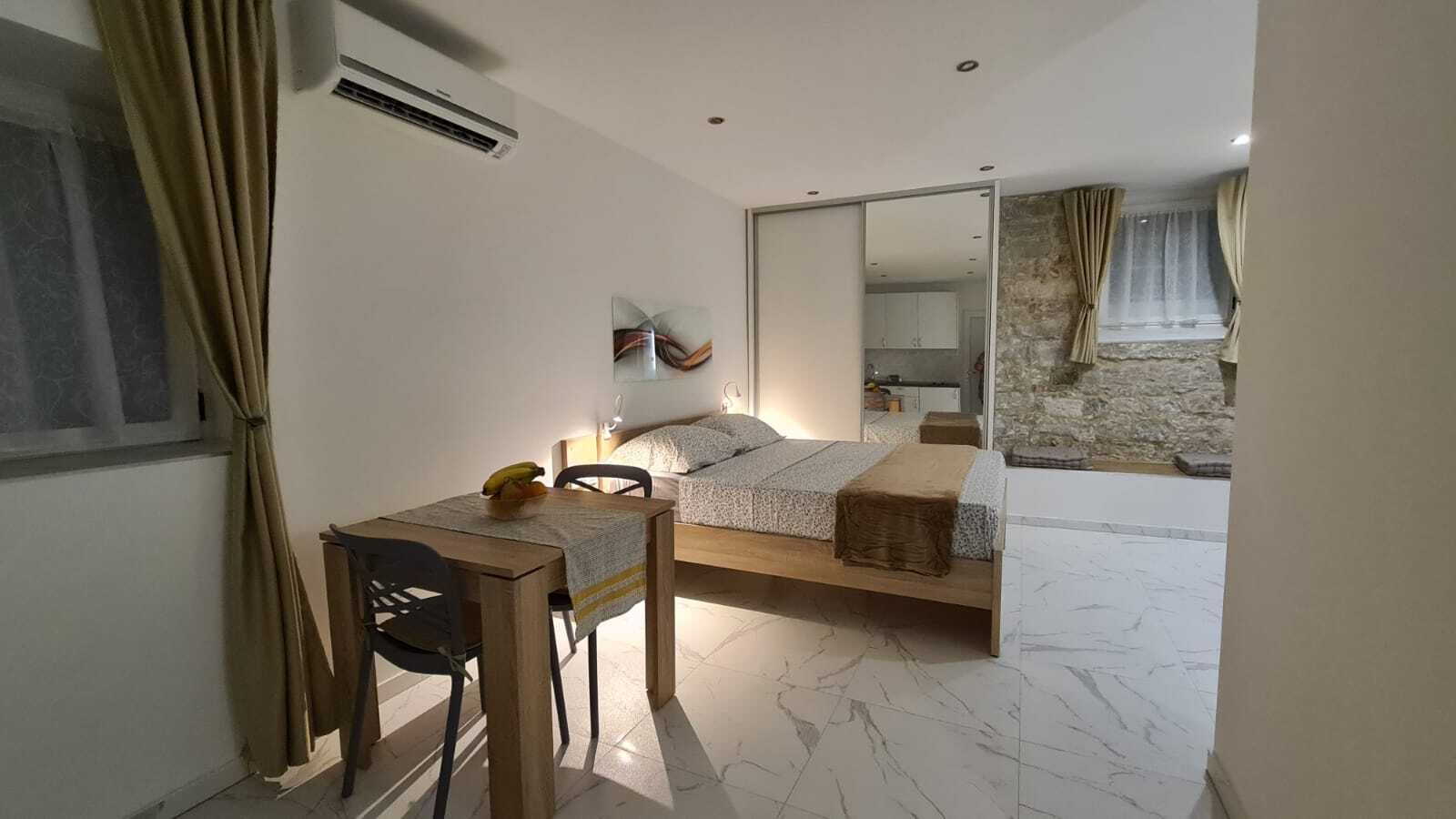A bedroom with a bed, a desk, and a chair. There is a window with curtains, an air conditioning unit, and a mirror on the wall.