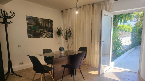 Een eethoek met een glazen tafel, zwarte stoelen, een potplant en een schilderij aan de muur.