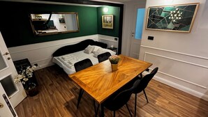 Een compacte kamer met een bed, een houten eettafel met stoelen, een klein bijzettafeltje en een spiegel aan de muur.